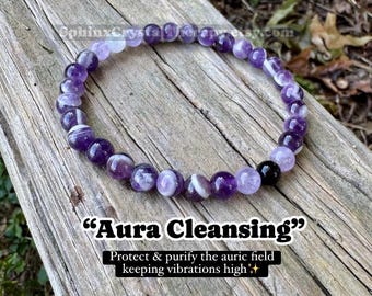 May include: A circular beaded bracelet featuring polished purple amethyst stones with white banding and one solid black bead. The text "Aura Cleansing" and "Protect & purify the auric field keeping vibrations high" is printed below the bracelet.