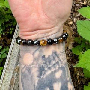 May include: A stretch bracelet featuring alternating round beads in polished black, metallic gray, and golden-brown tiger eye. The beads are strung on an elastic cord, creating a circular band that fits snugly around the wrist. The surface of the beads reflects light, highlighting the smooth texture and distinct color variations of the stones.