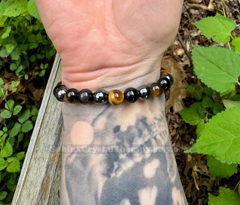May include: A stretch bracelet featuring alternating round beads in polished black, metallic gray, and golden-brown tiger eye. The beads are strung on an elastic cord, creating a circular band that fits snugly around the wrist. The surface of the beads reflects light, highlighting the smooth texture and distinct color variations of the stones.