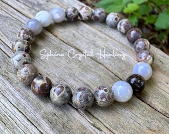 May include: A beaded bracelet featuring a variety of stone beads in shades of brown, gray, and white. The bracelet is resting on a wooden surface. The text "Sphinx Crystal Healing" is visible.