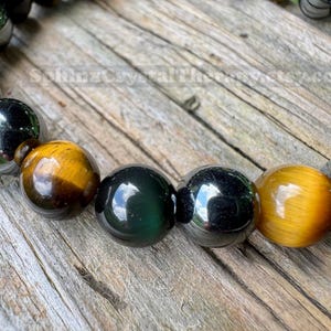 May include: A beaded bracelet featuring alternating polished spherical beads in golden-brown tiger eye, dark metallic hematite, and deep green obsidian. The beads are strung together in a repeating pattern, showcasing natural stone textures and reflective surfaces.