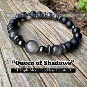 May include: A beaded bracelet featuring a mix of black, gray, and silver beads. The bracelet includes matte black, round beads, and larger, iridescent gray beads. Text on the image reads "Queen of Shadows" and "Triple Moon Goddess Hecate".