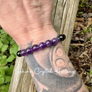 May include: A stretch bracelet featuring a series of smooth, round purple amethyst beads arranged in the center, flanked by two glossy black beads on each end. The beads are strung together on an elastic cord, creating a simple circular design.
