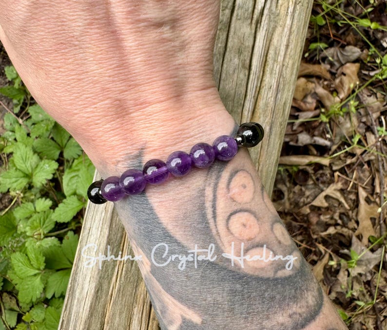 May include: A stretch bracelet featuring a series of smooth, round purple amethyst beads arranged in the center, flanked by two glossy black beads on each end. The beads are strung together on an elastic cord, creating a simple circular design.