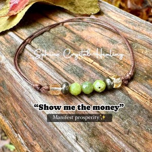 May include: A brown string bracelet with green and gold beads. The bracelet has the text "Show me the money" and "Manifest prosperity" on it.
