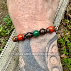 May include: A beaded bracelet featuring a mix of colorful gemstones. The bracelet includes red, brown, green, and smoky quartz beads, accented with silver-toned spacers. The bracelet is designed for everyday wear.