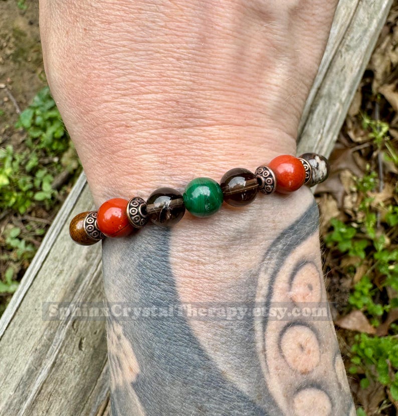 May include: A beaded bracelet featuring a mix of colorful gemstones. The bracelet includes red, brown, green, and smoky quartz beads, accented with silver-toned spacers. The bracelet is designed for everyday wear.