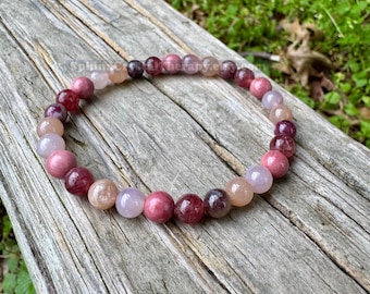 May include: A circular beaded bracelet featuring polished spherical stones in varying shades of pink, mauve, and deep burgundy. The beads are strung together in a continuous loop, displaying a smooth, glossy texture and natural color variations.