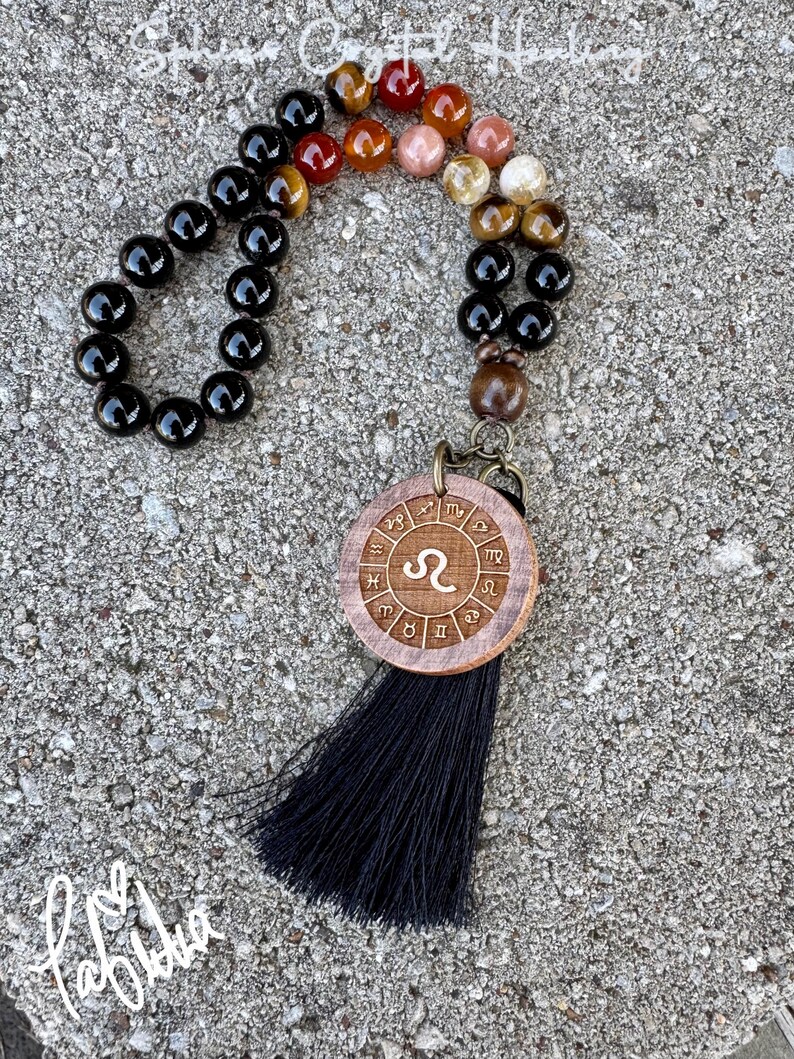 Puede incluir: Un collar de cuentas negras con un colgante de madera que presenta el s&iacute;mbolo astrol&oacute;gico de Leo y los 12 signos del zod&iacute;aco. El collar tiene una borla negra.