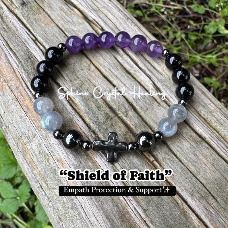 May include: A beaded bracelet featuring purple amethyst, black, and grey beads with a central black cross charm. The text "Sphinx Crystal Healing" and "Shield of Faith" with "Empath Protection & Support" is overlaid on the image of the bracelet.