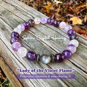 May include: A purple, pink, and white beaded bracelet with a grey stone charm. The bracelet is on a wooden surface and has the text "Sphinx Crystal Healing" and "Lady of the Violet Flame" Protection, intuition & aura clearing" on it.