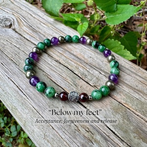 May include: A beaded bracelet featuring green, purple, and metallic grey spherical beads, with two dark red beads flanking a central silver-toned metal charm with an etched spiral design. The text "Below my feet" and "Acceptance, forgiveness and release" is visible.