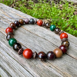 May include: A beaded bracelet featuring a variety of colorful gemstones. The bracelet includes red, brown, green, and black beads, with decorative metal accents. The bracelet is displayed on a weathered wooden surface.