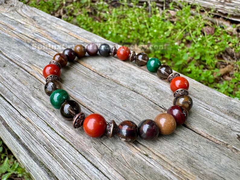 May include: A beaded bracelet featuring a variety of colorful gemstones. The bracelet includes red, brown, green, and black beads, with decorative metal accents. The bracelet is displayed on a weathered wooden surface.
