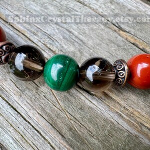 May include: Close-up of a beaded bracelet featuring a variety of gemstones. The bracelet includes red, green, and brown beads, separated by decorative metal spacers. The background is a weathered wooden surface.