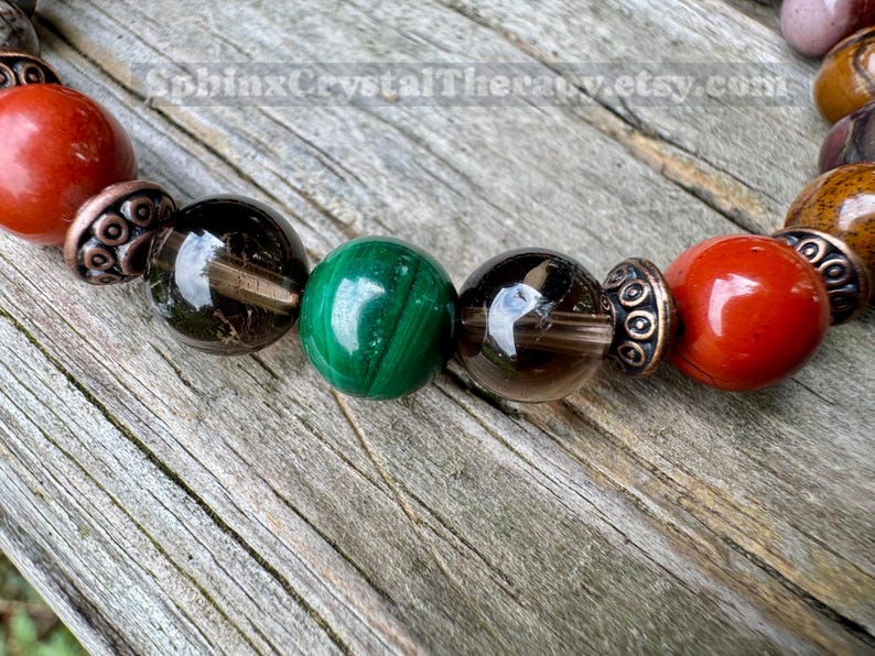 May include: Close-up of a beaded bracelet featuring a variety of gemstones. The bracelet includes red, green, and brown beads, separated by decorative metal spacers. The background is a weathered wooden surface.
