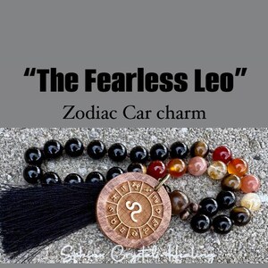 Puede incluir: Un abalorio de coche con borla negra que presenta un c&iacute;rculo de madera con el s&iacute;mbolo del zod&iacute;aco Leo y las palabras "The Fearless Leo" y "Zodiac Car charm".