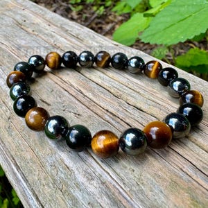 May include: A beaded bracelet featuring alternating polished round beads in shades of golden brown tiger eye and glossy black obsidian. The beads are strung together in a circular pattern, showcasing a smooth, reflective surface on each individual stone.