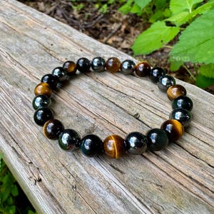 May include: A beaded bracelet featuring alternating round polished beads in shades of golden brown tiger's eye, glossy black obsidian, and metallic dark gray hematite, strung together on a flexible cord in a circular arrangement.