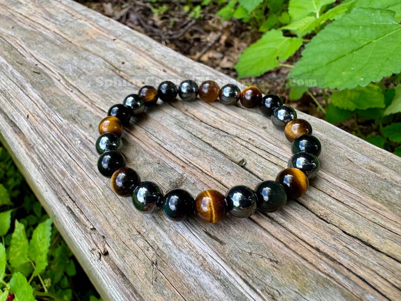 May include: A beaded bracelet featuring alternating round polished beads in shades of golden brown tiger's eye, glossy black obsidian, and metallic dark gray hematite, strung together on a flexible cord in a circular arrangement.