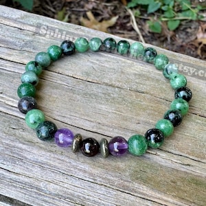 May include: A beaded bracelet featuring a mix of green, black, purple, and gold-colored beads. The bracelet is made of round beads and is displayed on a weathered wooden surface. The beads are strung together to form a complete circle.