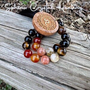 Puede incluir: Un amuleto de madera del zod&iacute;aco con el s&iacute;mbolo de Escorpio, rodeado de un brazalete de cuentas negras, marrones, naranjas y rojas.