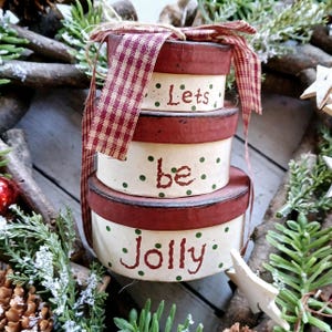 May include: A festive stack of three cream-coloured, round boxes with red rims and lids. The boxes are adorned with green polka dots and the words "Lets be Jolly" in a handwritten style. A red and white checked ribbon is tied around the boxes.