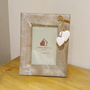 May include: A distressed, light grey rectangular photo frame. Two white heart-shaped ornaments hang from a twine bow. The frame has a visible logo: "The Home & Hearts Shop". The frame is suitable for a photograph.
