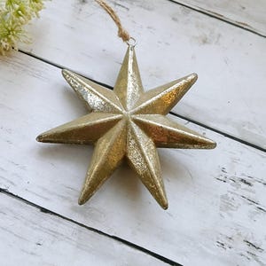 May include: A gold-coloured, eight-pointed star ornament with a rustic, textured finish. The star has a loop at the top for hanging.