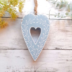 May include: A pale blue heart-shaped decoration with a heart-shaped void in the middle. It features white floral detailing around the edges. A jute string is attached for hanging. The background is a white wooden surface.
