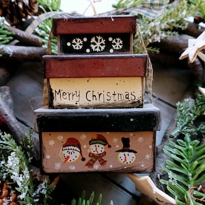 May include: A festive stack of three painted boxes. The top box is black with white snowflakes, the middle box reads "Merry Christmas", and the bottom box features snowman faces. The boxes are painted in shades of red, black, and beige.