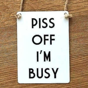 May include: White metal sign with black text that reads "Piss Off I'm Busy".