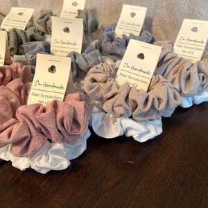 May include: Set of two handmade hair scrunchies in a light brown and a light pink color. The scrunchies are made of a soft, textured fabric and have a white elastic band.