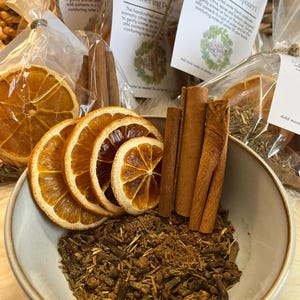 Può includere: Una ciotola di ceramica piena di pot-pourri invernale, con fette d'arancia essiccate, bastoncini di cannella e un mix di spezie aromatiche. L'immagine include etichette che recitano "Winter Blend Simmering Potpourri."