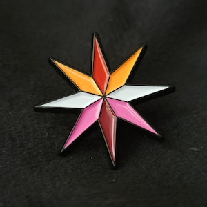 Puede incluir: Un pin de estrella de seis puntas con un contorno negro y un diseño colorido. La estrella está dividida en seis secciones, cada una de un color diferente: rojo, naranja, amarillo, blanco, rosa y granate.