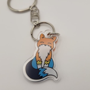 Puede incluir: Un llavero de acrílico transparente con un diseño de zorro de dibujos animados. El zorro es naranja y blanco con una cola azul y negra. El llavero está unido a un anillo de metal plateado.