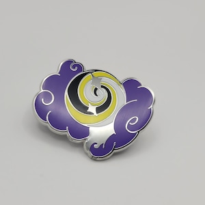 Puede incluir: Un pin de esmalte morado con un diseño estilizado de luna y estrellas. La luna es amarilla, negra y blanca, y las estrellas son blancas.