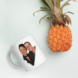 May include: White ceramic mug with a cartoon illustration of two people with brown skin, smiling and looking at the camera. The person on the left is wearing a black leather jacket and the person on the right is wearing a black shirt.