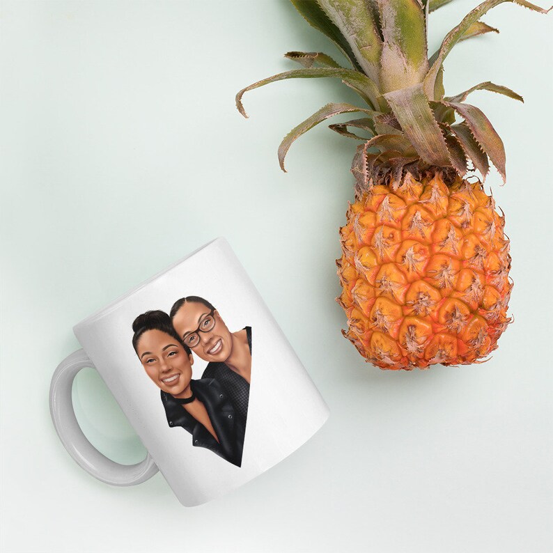 May include: White ceramic mug with a cartoon illustration of two people with brown skin, smiling and looking at the camera. The person on the left is wearing a black leather jacket and the person on the right is wearing a black shirt.