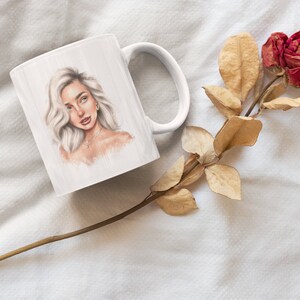 May include: White ceramic mug with a drawing of a woman with blonde hair and a gold necklace. The mug is on a white surface with a dried rose.