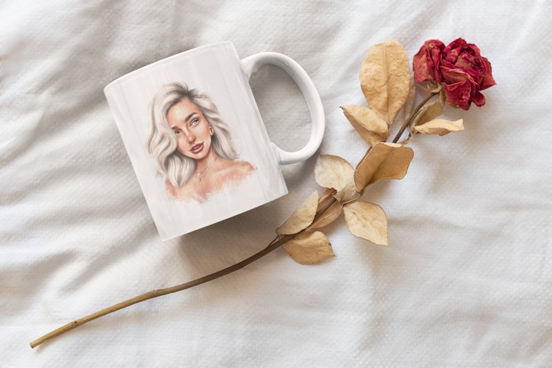 May include: White ceramic mug with a drawing of a woman with blonde hair and a gold necklace. The mug is on a white surface with a dried rose.