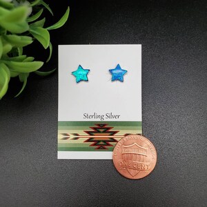 May include: A pair of sterling silver star earrings with blue opal stones. The earrings are displayed on a white card with the words "Sterling Silver" printed on it. A penny is included for size reference.