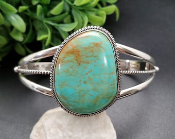 May include: Silver cuff bracelet featuring a large, teardrop-shaped turquoise stone with brown veining. The stone is set in a detailed, rope-like bezel. The bracelet has a split shank design.