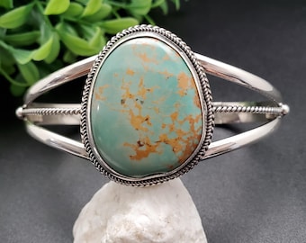 May include: A silver cuff bracelet featuring a large, oval turquoise stone with brown matrix. The stone is set in a decorative bezel with a twisted rope border. The bracelet has a split shank design.