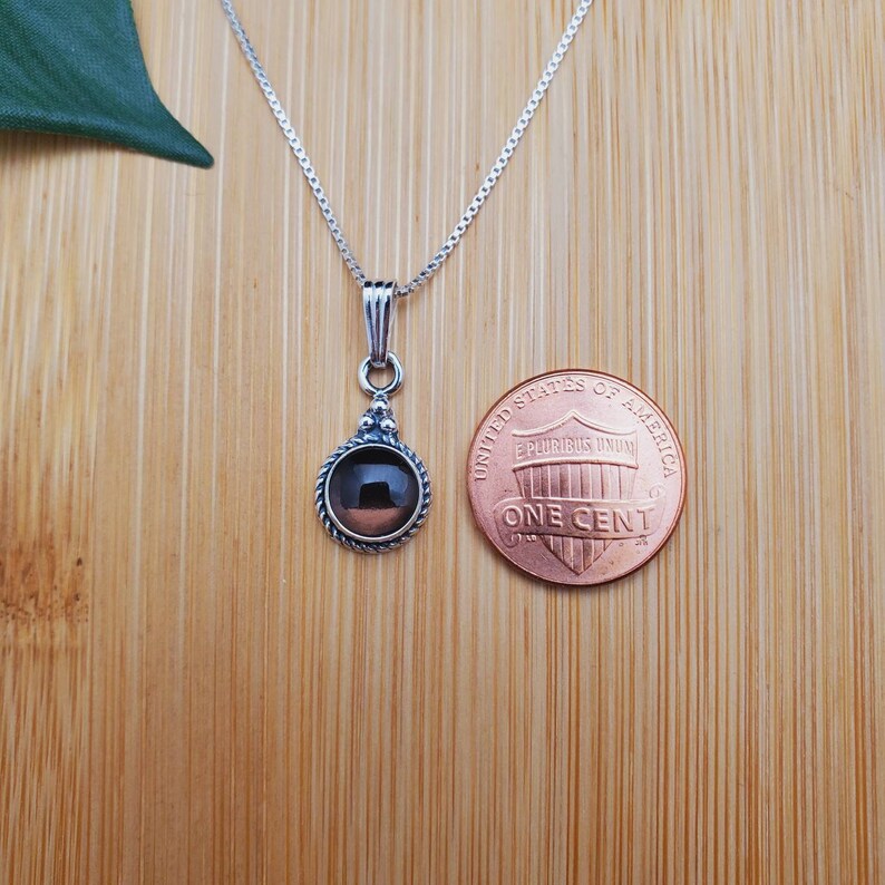 Pu&ograve; includere: Una collana con catena d'argento con un ciondolo che presenta una pietra preziosa rotonda di colore marrone scuro incastonata in una ghiera d'argento. Il ciondolo &egrave; circondato da un bordo d'argento con un design decorativo. La collana &egrave; mostrata accanto a un centesimo di dollaro americano per il confronto delle dimensioni.