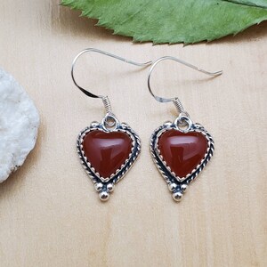 May include: A pair of silver heart-shaped earrings with red carnelian gemstones. The earrings have a delicate, ornate design with a beaded detail at the bottom.
