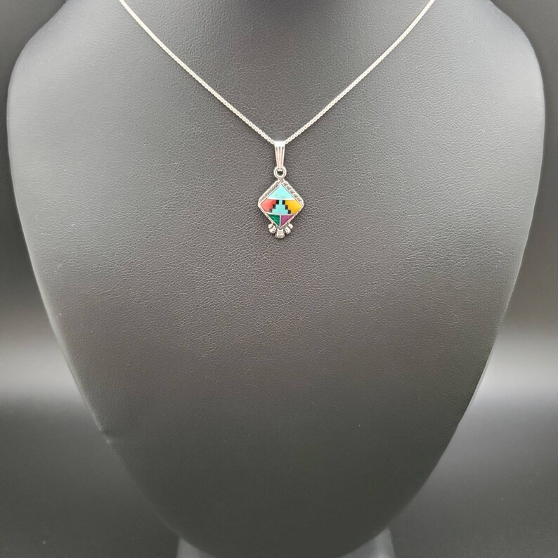 May include: A silver chain necklace with a small, square pendant featuring a geometric design of turquoise, red, yellow, green, and black stones.