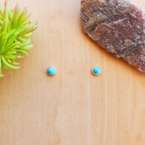 May include: A pair of turquoise stud earrings with silver settings. The earrings are displayed on a wooden surface.