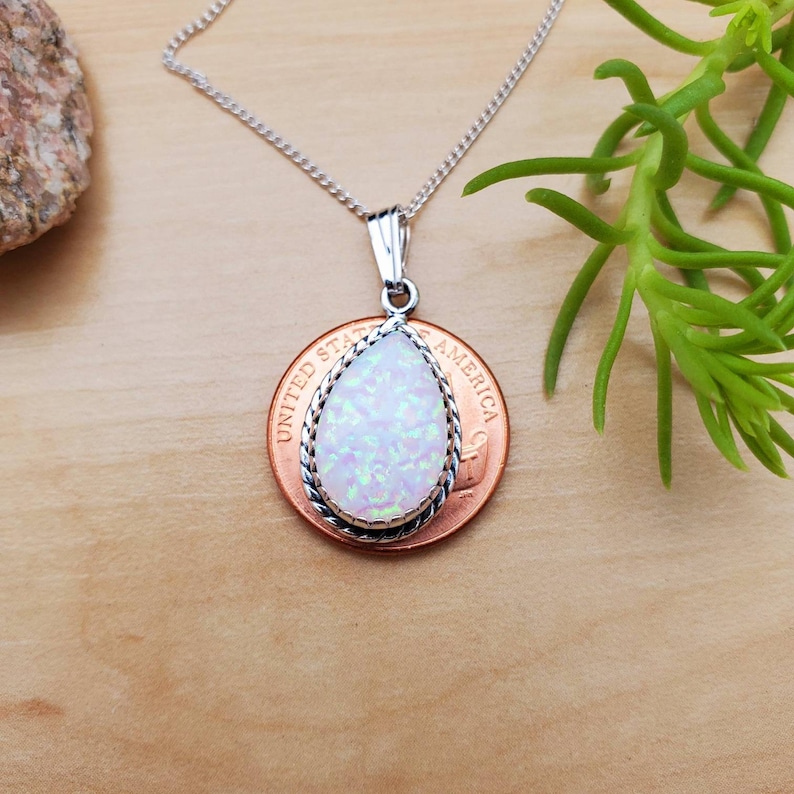 May include: A silver chain necklace with a teardrop-shaped white opal pendant. The pendant is set in a silver bezel with a rope-like border. The pendant is shown on top of a US penny.