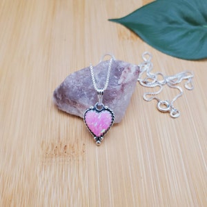 May include: A silver heart-shaped pendant necklace with a pink opal stone. The necklace is on a wooden surface with a small rock and a green leaf.